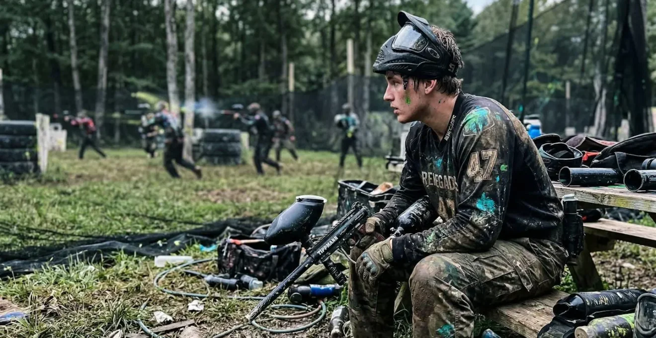 Un participant seul au bord d'un terrain de paintball, masque relevé, observe les autres jouer sans lui